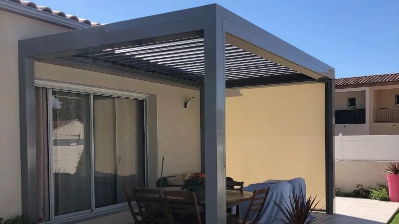 Moderne Aluminium Pergola mit Lamellendach auf Terrasse neben Haus und Gartenmöbeln.